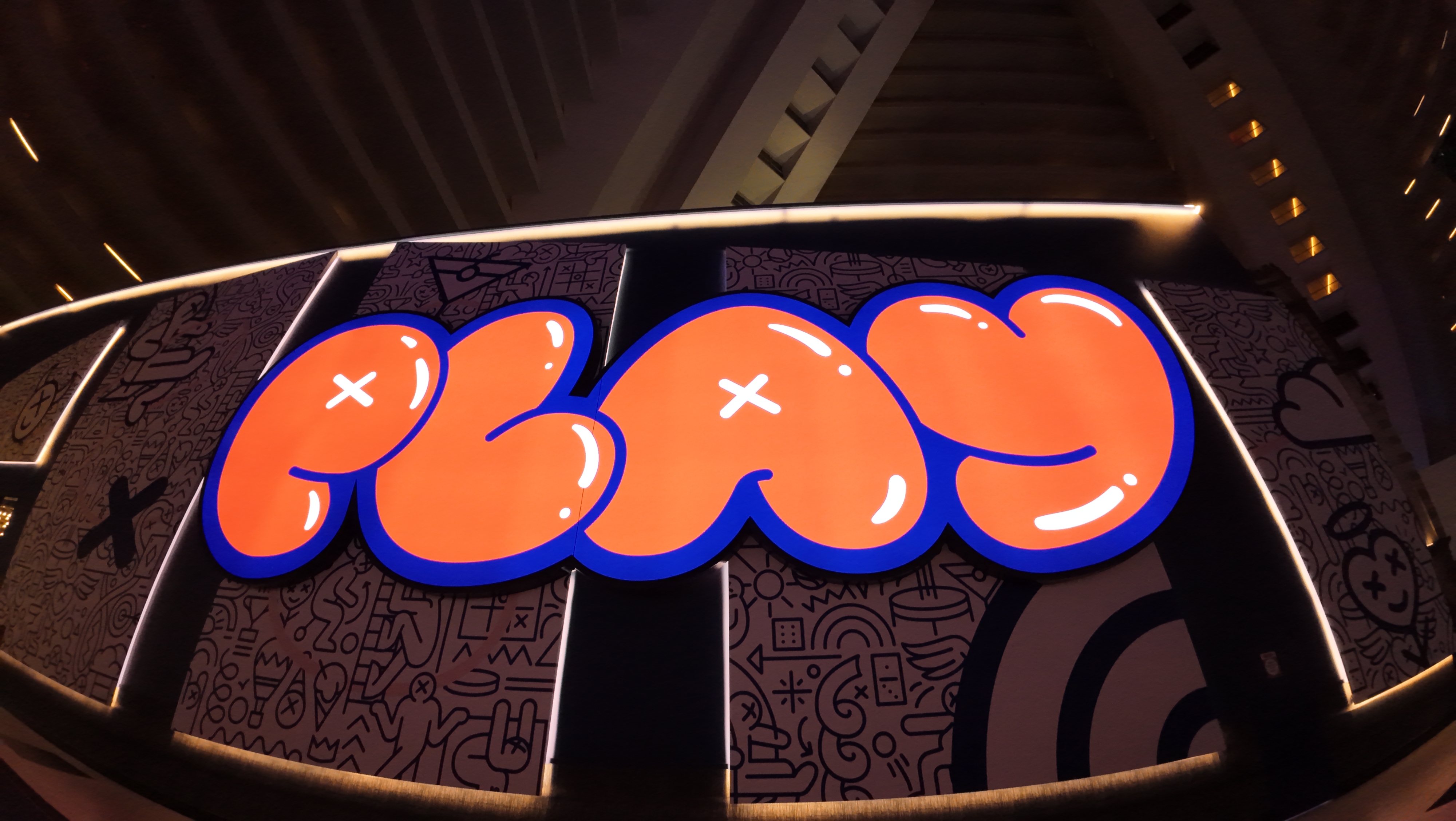  A large illuminated sign with bold orange letters sits in front of a black‑and‑white wall filled with graphic doodles and symbols.